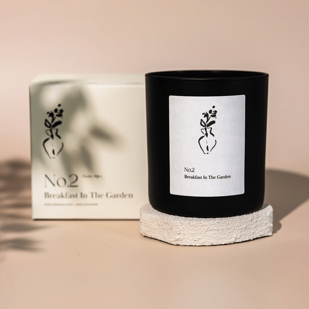 No. 2 Breakfast In The Garden (Swiss Alps) Candle | Lucky Owl in a black glass container with minimalist white label featuring a floral illustration, placed on a textured white stone coaster with matching product box in the background.