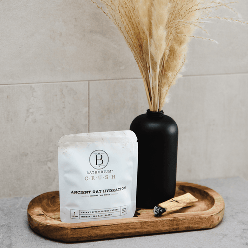 Ancient Oat Hydration Crush | Bathorium bath soak packet displayed on a wooden tray next to a black vase with dried pampas grass, highlighting skincare and wellness body care.