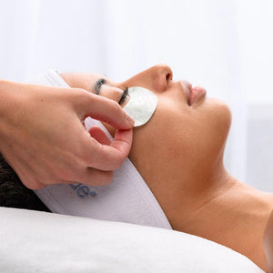 Close-up of a woman lying down with a calm expression as a hand gently applies a PolyPeptide Collagel+ Eye Mask | HydroPeptide under her eye during a skincare treatment.