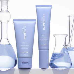 Two light purple HydroPeptide skincare tubes labeled PEEL 1 and PEEL 2 from the Polish & Plump Face Peel Kit | HydroPeptide surrounded by clear glass lab beakers with blue liquid, emphasizing skincare and wellness supplements.
