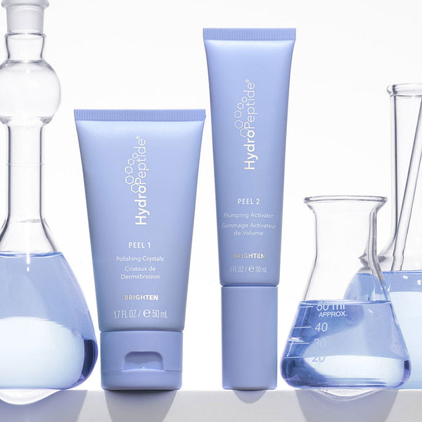 Two light purple HydroPeptide skincare tubes labeled PEEL 1 and PEEL 2 from the Polish & Plump Face Peel Kit | HydroPeptide surrounded by clear glass lab beakers with blue liquid, emphasizing skincare and wellness supplements.