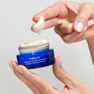 Close-up of a hand scooping creamy moisturizer from a blue jar labeled Power Lift | HydroPeptide, highlighting luxurious skincare for hydration and lifting benefits.