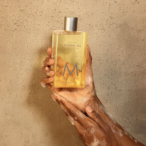 Hand holding a bottle of Shower Gel | Moroccanoil with wet, soapy hands against a sandy beige background, highlighting skincare and body care wellness.