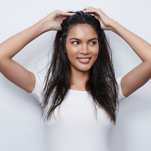 Smiling woman with long dark hair applying Volumizing Mousse | Moroccanoil, demonstrating a hair care routine for natural volume and smooth texture.