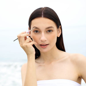 Woman with long dark hair applying makeup to her eyebrows using Eyebrow Pencil | Chella against a bright, softly blurred outdoor background.