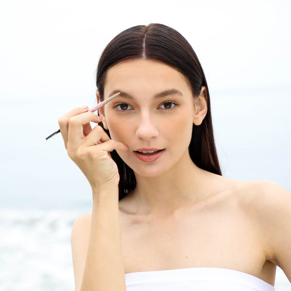 Woman with long dark hair applying makeup to her eyebrows using Eyebrow Pencil | Chella against a bright, softly blurred outdoor background.