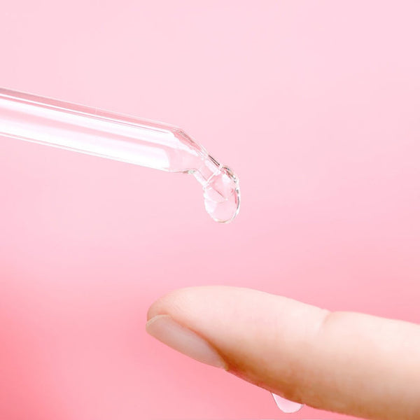Close-up of a dropper dispensing a clear serum drop onto a fingertip with a soft pink background, highlighting Daily Dose Of Water™ Hyaluronic Acid Hydrating Serum | ClarityRx for skincare.