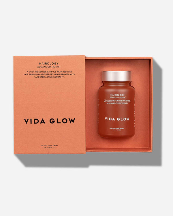 Open box showing a brown bottle with silver lid labeled Hairology Capsules | VIDA GLOW inside an orange presentation box designed for wellness supplements supporting hair growth and repair.