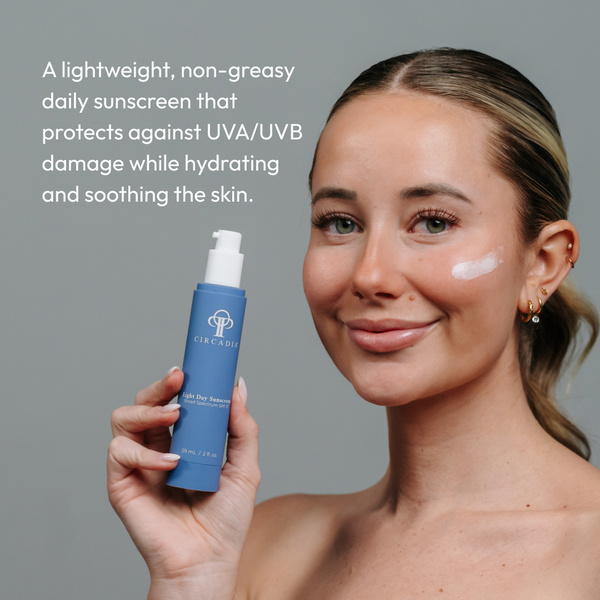 Woman with clear skin holding a blue bottle of Light Day Sunscreen Broad Spectrum SPF 37 | Circadia, with a small amount of sunscreen applied on her cheek against a neutral background.