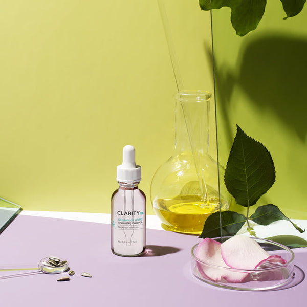 Glimmer Of Hope™ Shimmering Facial Oil | ClarityRx bottle displayed alongside a glass beaker with golden oil, a petri dish with pink flower petals, and green leaves on a pastel background, highlighting skincare and wellness.