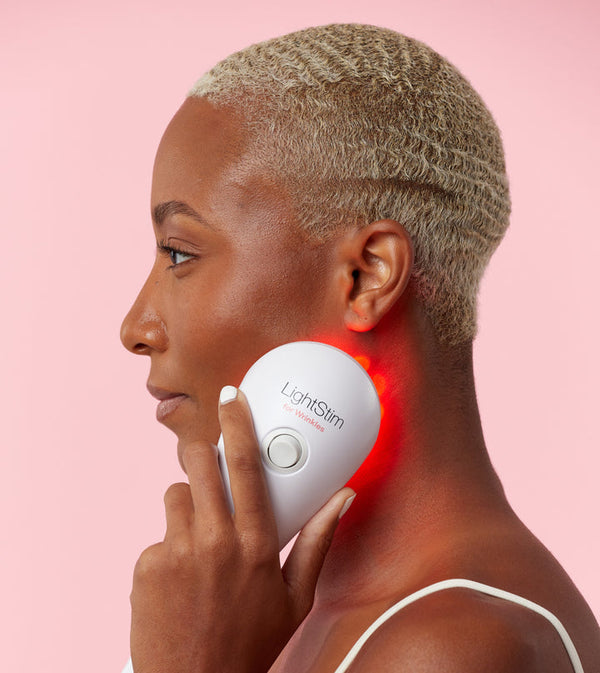 Woman using the LightStim For Wrinkles | LightStim device emitting red light on her jawline against a pink background for skincare and wellness care.