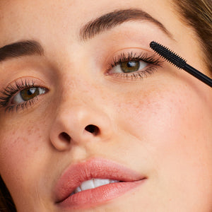 Close-up of a woman applying Lash Fixation Length & Definition Tubing Mascara | Jane Iredale to her upper eyelashes, showcasing natural makeup with defined lashes and clear skin.