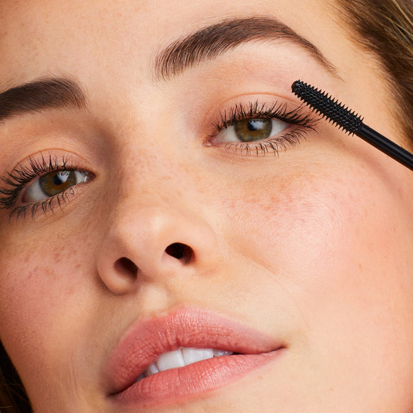 Close-up of a woman applying Lash Fixation Length & Definition Tubing Mascara | Jane Iredale to her upper eyelashes, showcasing natural makeup with defined lashes and clear skin.