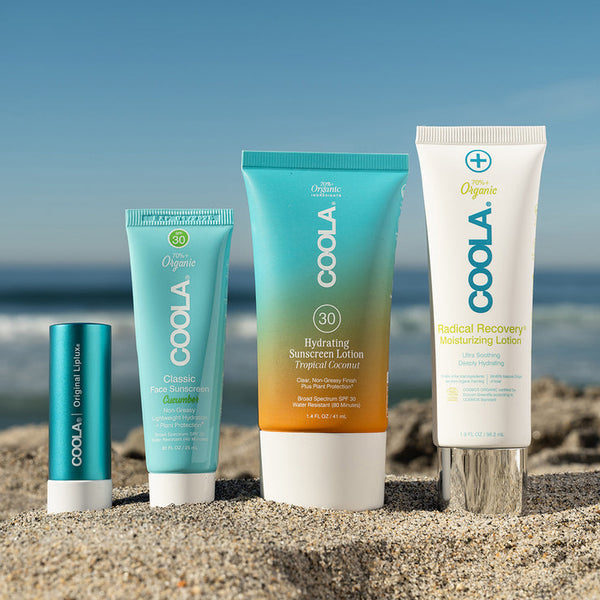 Four COOLA skincare products from the Suncare Travel Essentials Set | COOLA displayed on sand at the beach, highlighting sunscreen and moisturizing lotion for sun protection and hydration.