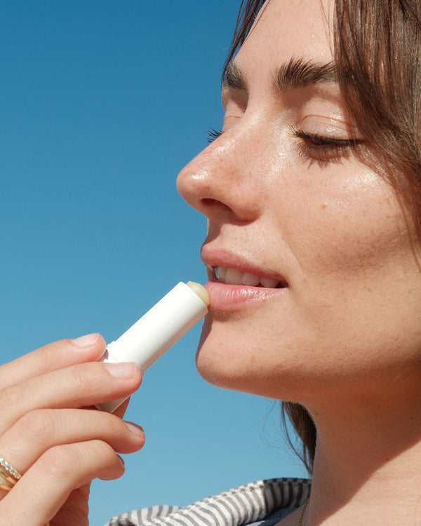 Close-up of a woman applying lip balm outdoors under a clear blue sky, showcasing the Suncare Travel Essentials Set | COOLA for skincare and sun protection.