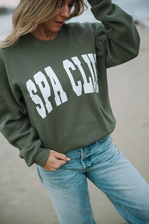 Woman outdoors wearing an olive green Spa Club Crew Neck Sweatshirt | Lucky Owl with white lettering paired with light blue jeans, styled casually for wellness and comfort.