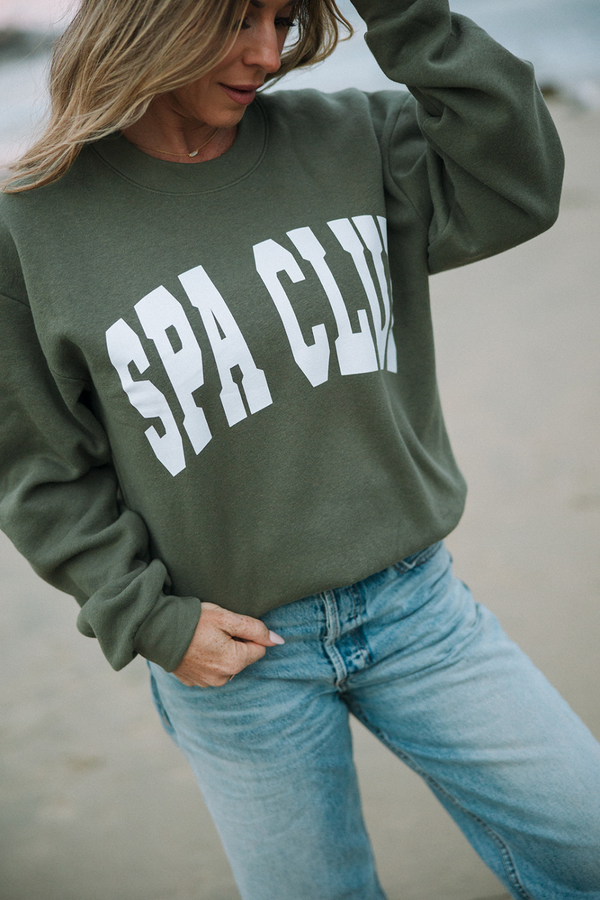 Woman outdoors wearing an olive green Spa Club Crew Neck Sweatshirt | Lucky Owl with white lettering paired with light blue jeans, styled casually for wellness and comfort.