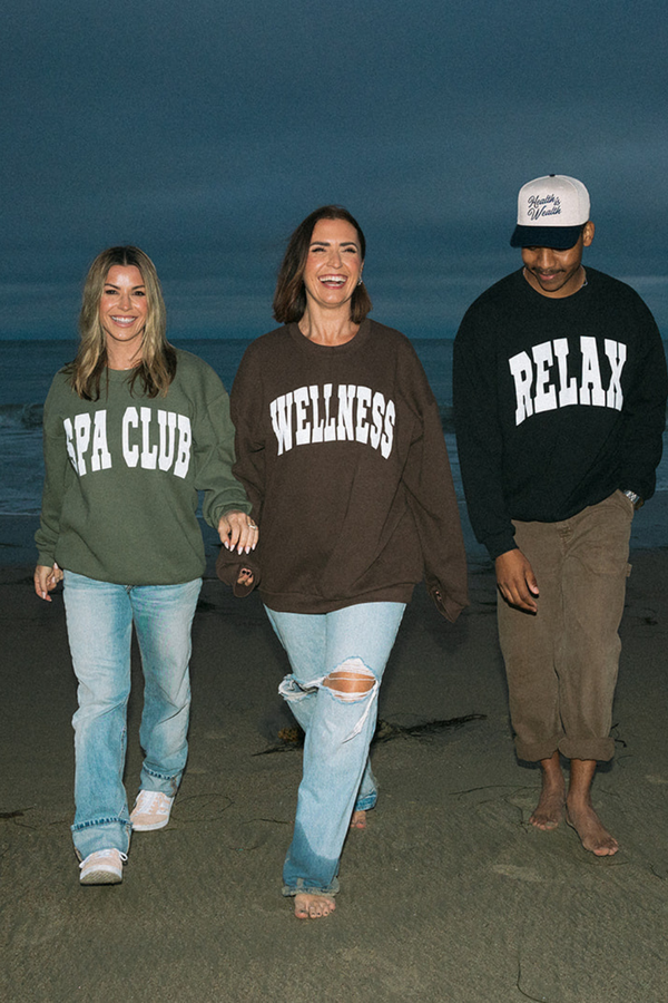 Three people walking on a beach at dusk wearing casual sweatshirts, one wearing the Spa Club Crew Neck Sweatshirt | Lucky Owl in olive green paired with light blue jeans.