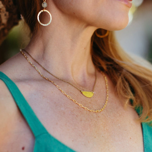 Close-up of a woman wearing layered gold necklaces and a textured gold hoop earring, showcasing the Frolic Paperclip Necklace /silver tone | Purpose Jewelry against a green top.