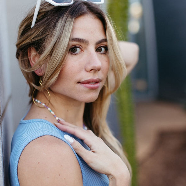 Close-up of a woman wearing Olivia Hoop Earrings | Purpose Jewelry with a casual blue top, natural makeup, and a colorful beaded necklace in an outdoor setting.