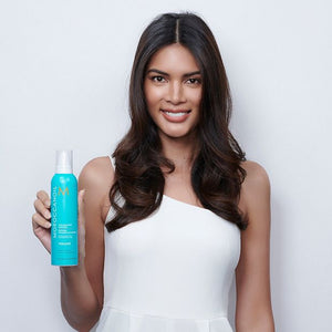 Smiling woman with long wavy hair wearing a white sleeveless dress holding a blue bottle of Volumizing Mousse | Moroccanoil against a plain light background.