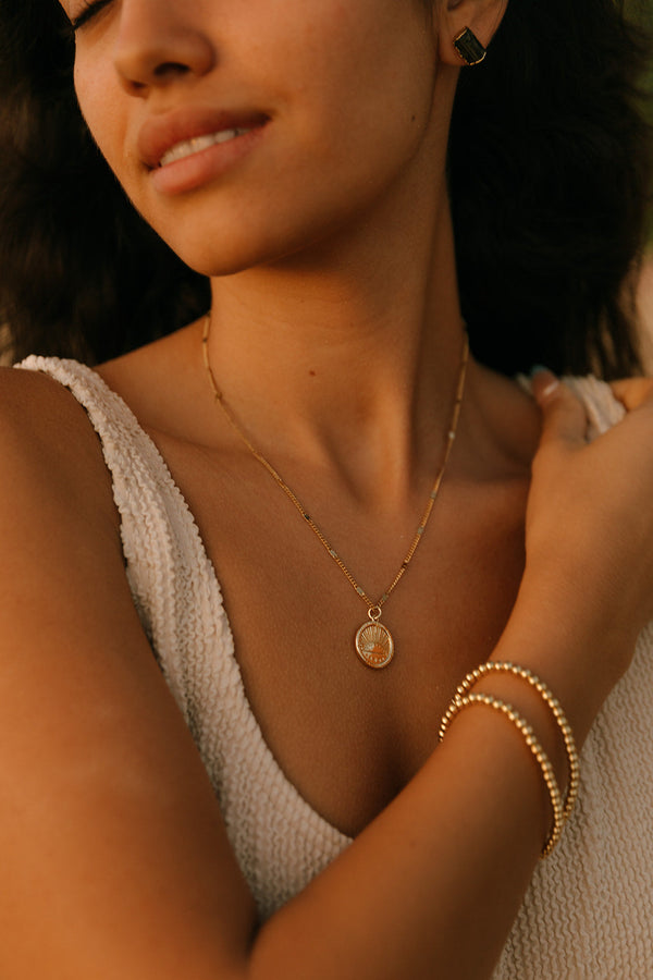 Close-up of a woman wearing a delicate gold pendant necklace with a sunburst design and matching gold bracelets, showcasing the Golden Sunrise Necklace | Meghan Bo.