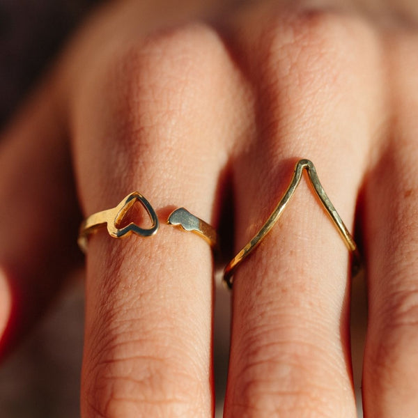 Close-up of two gold rings worn on fingers, one shaped like an open heart and the other featuring a minimalist chevron design, showcasing the Journey Ring | Purpose Jewelry.