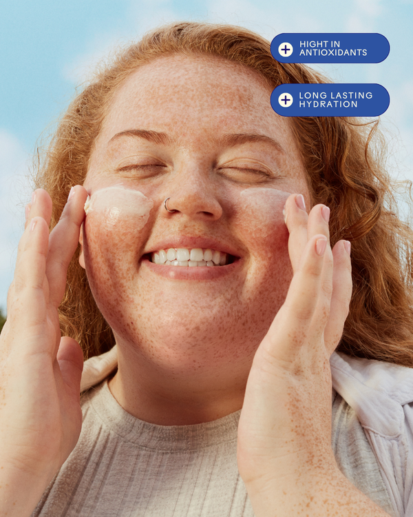 Happy woman with freckles applying moisturizing cream to her cheeks outdoors, highlighting the benefits of Radiance Grape Stem Cell + Squalane Ultra-Hydrating Cream | Three Ships for skincare hydration and antioxidants.