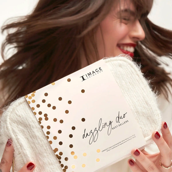 Woman with red nail polish holding a white textured skincare product labeled Dazzling Duo | IMAGE Skincare with gold confetti design, smiling joyfully in a bright setting.