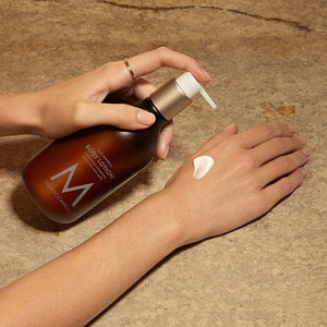 Hand applying creamy Body Lotion | Moroccanoil from a brown pump bottle onto the back of the other hand on a textured stone surface, showcasing skincare body care.