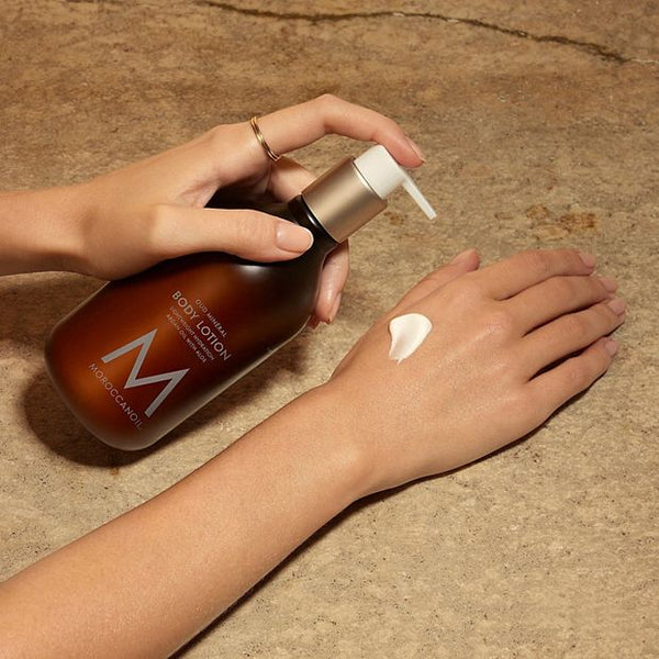 Hand applying creamy Body Lotion | Moroccanoil from a brown pump bottle onto the back of the other hand on a textured stone surface, showcasing skincare body care.