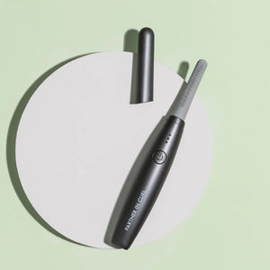 Deluxe Heated Eyelash Curler | Chella in sleek black design resting on a light circular platform with a minimal pale green background, highlighting its compact and modern wellness device style.