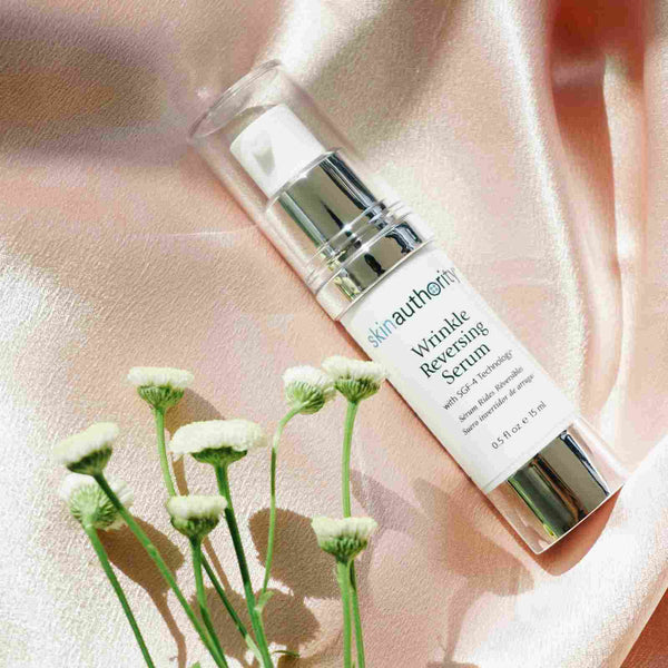 Complete Serum | Skin Authority bottle with a pump dispenser placed on a soft peach satin fabric next to small white flowers, highlighting skincare and wellness themes.