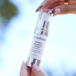 Hands holding a clear pump bottle of Complete Serum | Skin Authority skincare product against a blurred natural outdoor background.