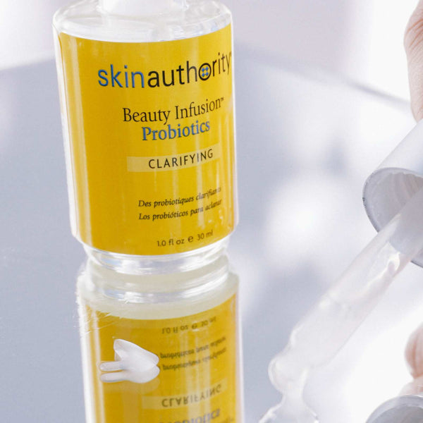 Close-up of a clear bottle of Beauty Infusion Probiotics for Clarifying | Skin Authority with yellow labeling, next to a hand holding a dropper dispensing clear serum on a reflective surface.