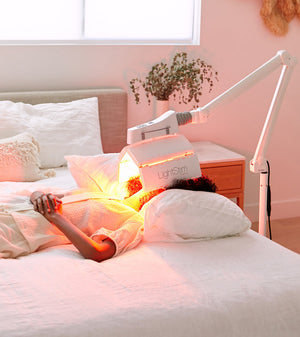 Person lying on a bed receiving red light therapy on their face from a LightStim ProPanel | LightStim device mounted on an adjustable arm in a cozy bedroom setting