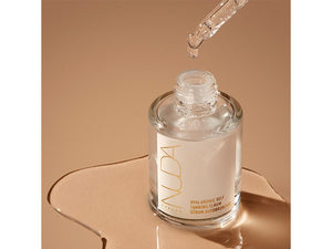 Clear glass bottle of Hyaluronic Self Tanning Serum | NUDA with dropper releasing serum, displayed on a smooth beige surface, highlighting skincare and body care wellness.