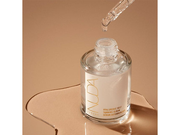 Clear glass bottle of Hyaluronic Self Tanning Serum | NUDA with dropper releasing serum, displayed on a smooth beige surface, highlighting skincare and body care wellness.