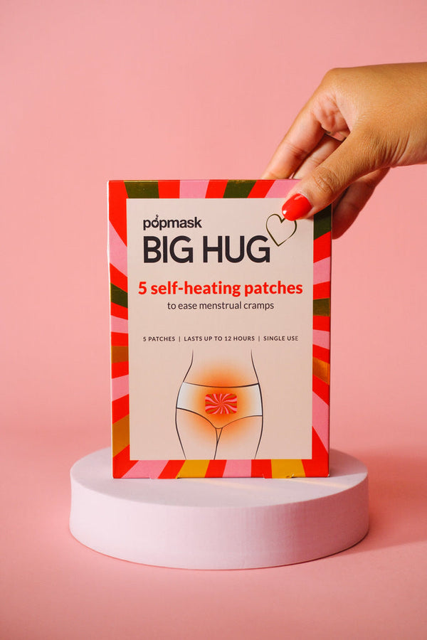 Hand holding a box of Big Hug - 5 Pack | Popmask self-heating patches designed to ease menstrual cramps, displayed on a round pink pedestal against a pink background.