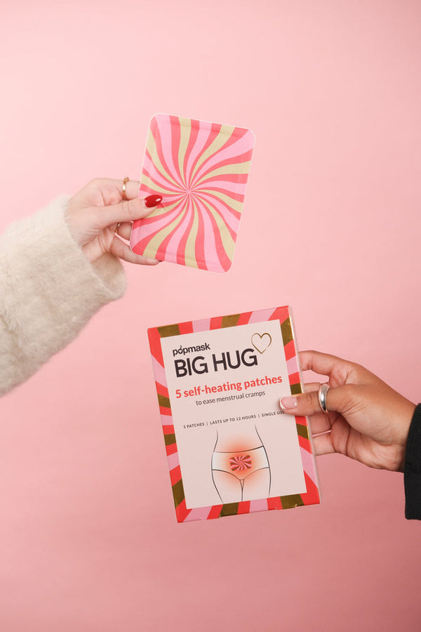 Hands holding a colorful packaging and a pink and red swirled patch for Big Hug - 5 Pack | Popmask self-heating patches designed for menstrual cramp relief on a soft pink background.