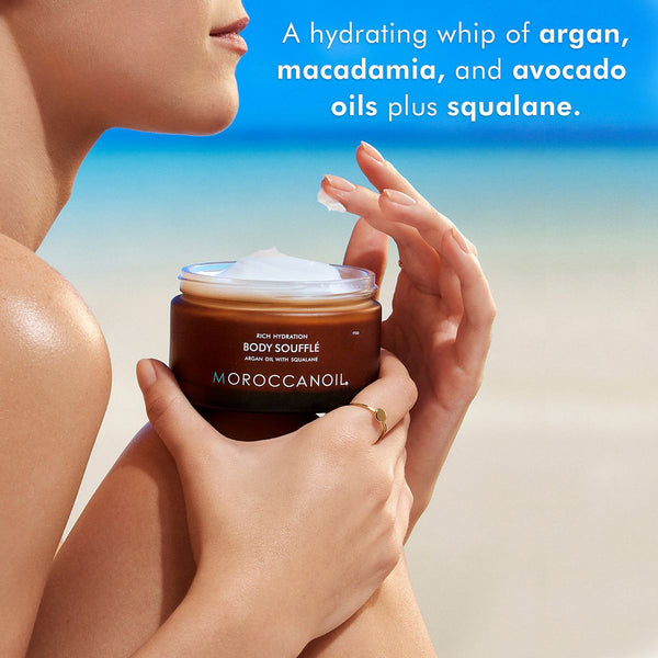 Person holding a jar of Body Soufflé | Moroccanoil against a beach backdrop, highlighting hydrating ingredients like argan, macadamia, avocado oils, and squalane for skincare.