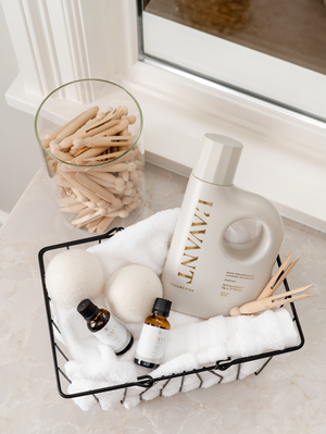 Wool Dryer Balls | L'AVANT Collective arranged in a wire basket with white towels, a white detergent bottle, two small amber bottles, wooden clothespins, and a glass jar filled with wooden clothespins on a marble surface.