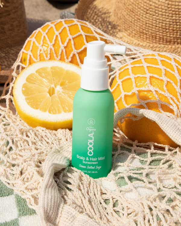 Scalp & Hair Mist Organic Sunscreen SPF 30 | COOLA bottle with a white pump on a beige netted bag, surrounded by fresh lemons and oranges in natural sunlight.