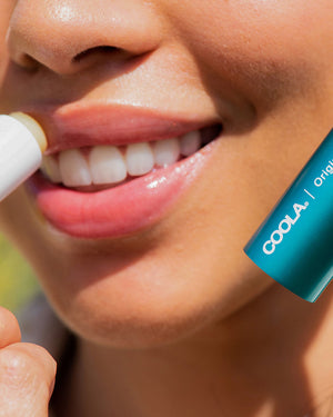 Close-up of a person smiling while applying Classic Liplux® Organic Lip Balm Sunscreen SPF 30 - Original | COOLA to their lips for natural skincare and sun protection.