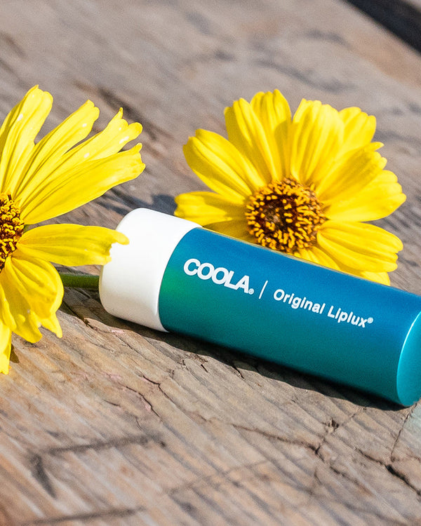 Classic Liplux® Organic Lip Balm Sunscreen SPF 30 - Original | COOLA lip balm tube next to bright yellow flowers on rustic wooden surface, emphasizing natural skincare and wellness.