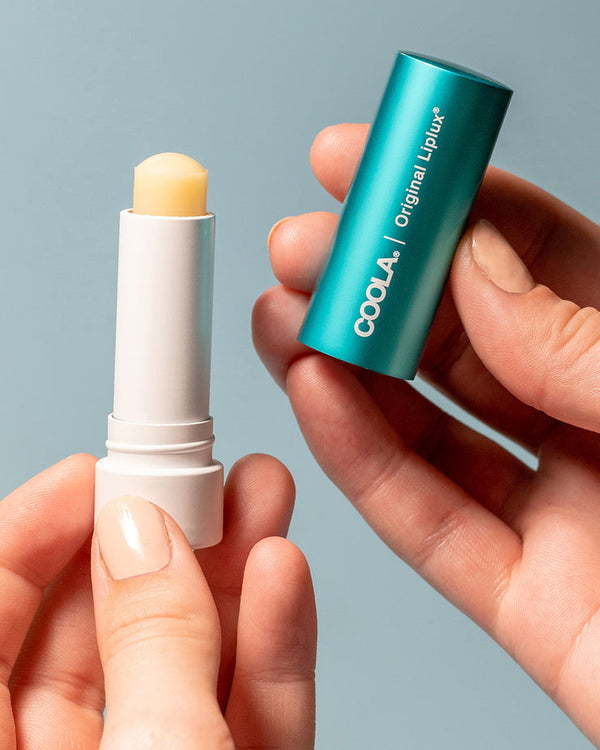 Hands holding Classic Liplux® Organic Lip Balm Sunscreen SPF 30 - Original | COOLA with the cap removed to show the balm inside against a plain blue background.