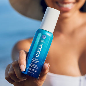 Hand holding a blue bottle of Classic Face Sunscreen Mist SPF 50 | COOLA with a white cap, shown outdoors with a blurred background and a smiling person.
