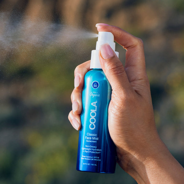 Hand holding and spraying Classic Face Sunscreen Mist SPF 50 | COOLA in a blue bottle, demonstrating lightweight skincare and sun protection with a fine mist spray outdoors.