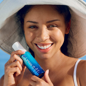 Woman wearing a sun hat smiling and holding a blue bottle of Classic Face Sunscreen Mist SPF 50 | COOLA against a clear sky background, promoting skincare and sun protection.