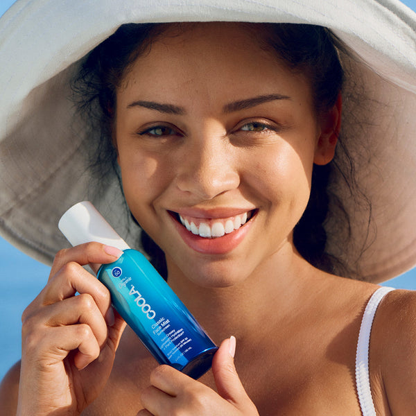 Woman wearing a sun hat smiling and holding a blue bottle of Classic Face Sunscreen Mist SPF 50 | COOLA against a clear sky background, promoting skincare and sun protection.